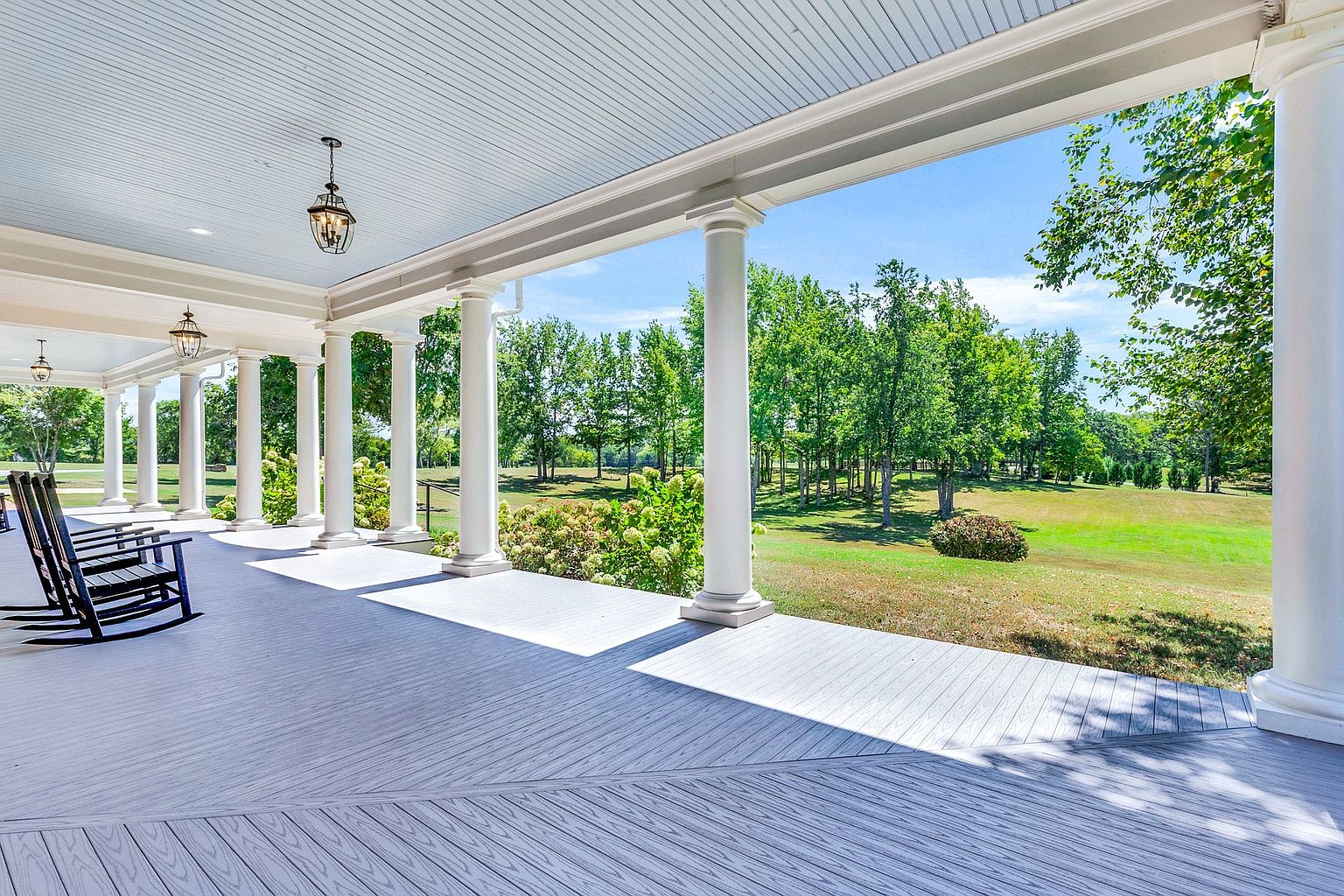 This expansive, covered wrap-around porch features classic white columns and a light-colored composite deck, offering a serene vantage point overlooking a lush, green landscape. Two black rocking chairs are positioned to take in the view, creating an inviting and peaceful atmosphere. The perspective is wide and cinematic, emphasizing the home's connection to the surrounding natural beauty.