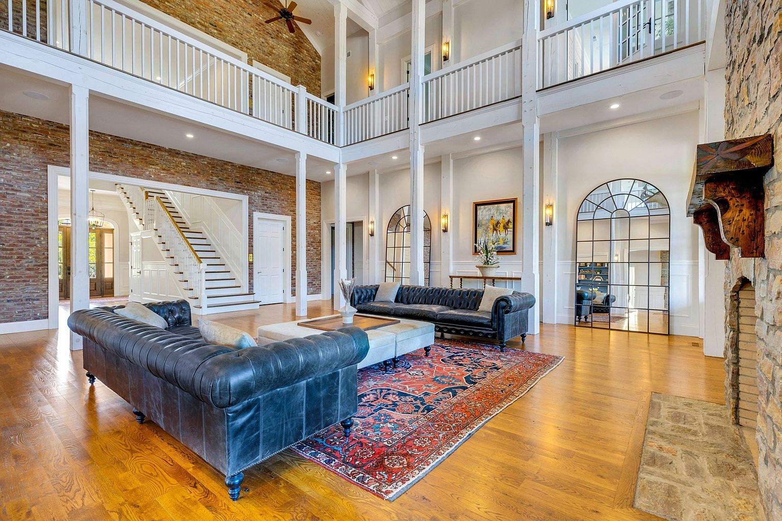 This grand, double-height living room features a striking blend of rustic brick and stone elements paired with elegant white architectural columns and railings. The space is anchored by two luxurious dark leather tufted sofas and a large, ornate area rug, creating a sophisticated yet inviting atmosphere. The perspective captures the expansive scale of the room, highlighting the dramatic staircase in the background and the arched floor-to-ceiling mirror that enhances the sense of openness.