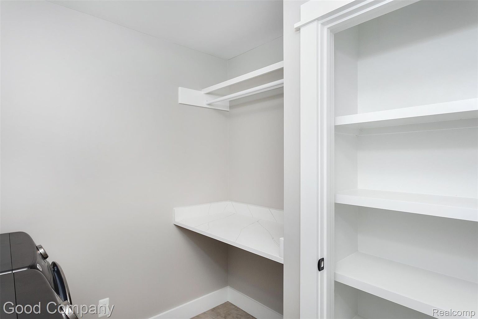 This is a well-organized laundry room featuring a countertop for folding, a hanging rod for clothes, and built-in shelving for storage. The color palette is neutral, with light gray walls and white trim, creating a clean and functional space. A washing machine is partially visible on the left.