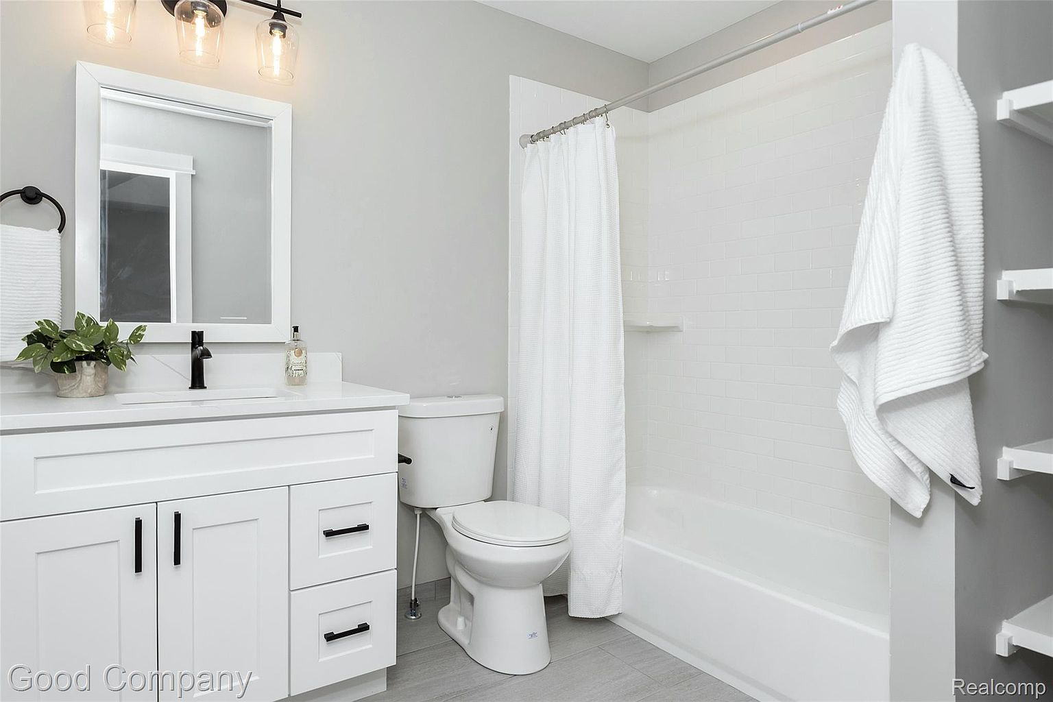 This is a well-lit bathroom featuring a white vanity with a black faucet and a framed mirror above. A toilet is positioned next to the vanity, and a bathtub with a white shower curtain is visible in the background. The bathroom has a clean and modern aesthetic, with gray flooring and light gray walls.