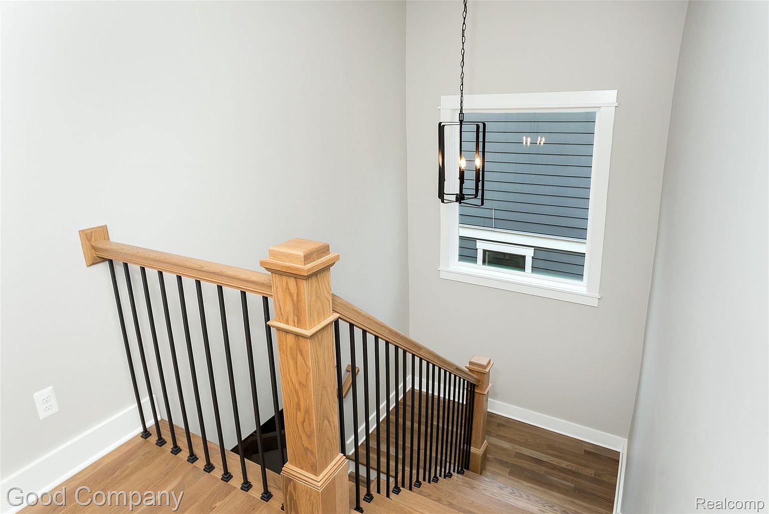 This image showcases a well-lit interior staircase with wooden steps and a black metal railing. A modern, rectangular pendant light hangs near a window, which provides natural light and a view of the exterior. The walls are painted in a neutral tone, creating a clean and inviting atmosphere.