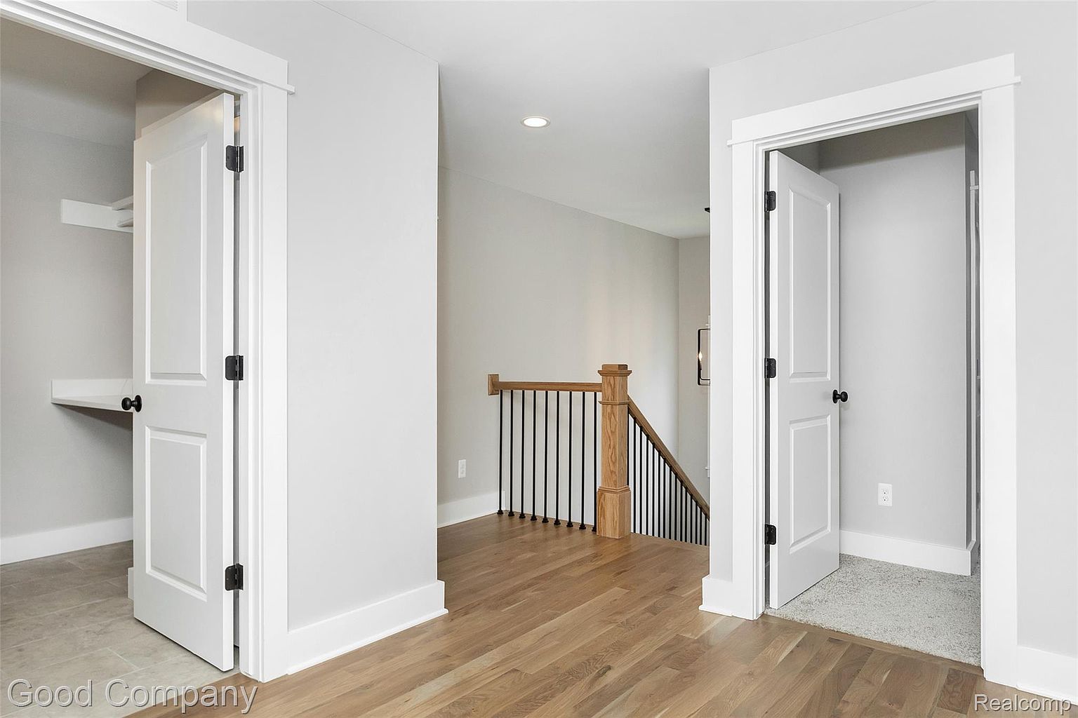 This interior shot showcases a hallway with hardwood flooring and white trim. Two open doorways reveal adjacent rooms, one with a built-in desk and the other with carpet. A staircase with black iron railings and a light wood banister is visible in the background, adding depth to the scene.