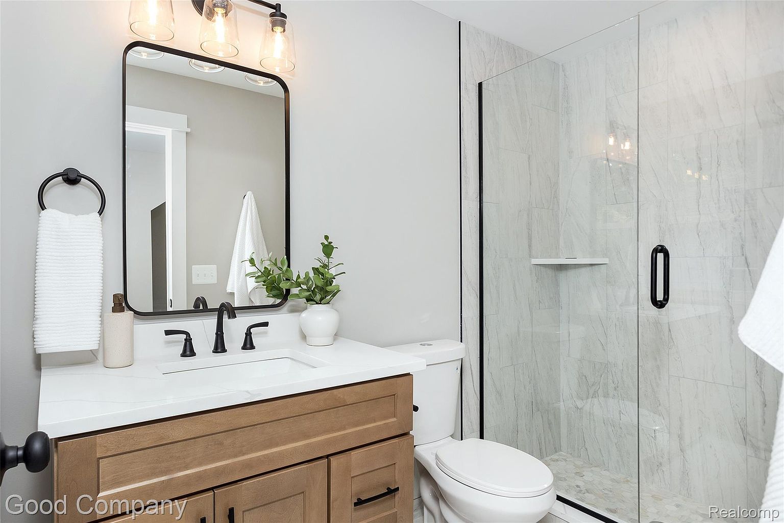 This is a well-lit bathroom featuring a modern design with a wood-toned vanity, white countertop, and black fixtures. A black-framed mirror hangs above the sink, and a glass-enclosed shower is visible to the right. The color palette is neutral, creating a clean and inviting space.