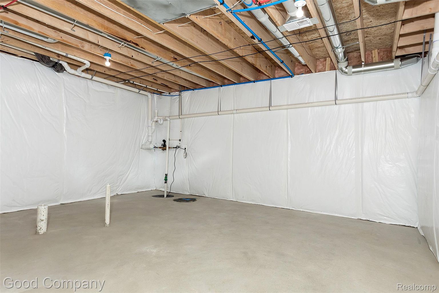 This is an interior shot of a basement, featuring a concrete floor and white plastic-covered walls. The ceiling is exposed with wooden beams and various pipes and wiring. The space appears unfinished but clean, offering a blank canvas for potential customization.