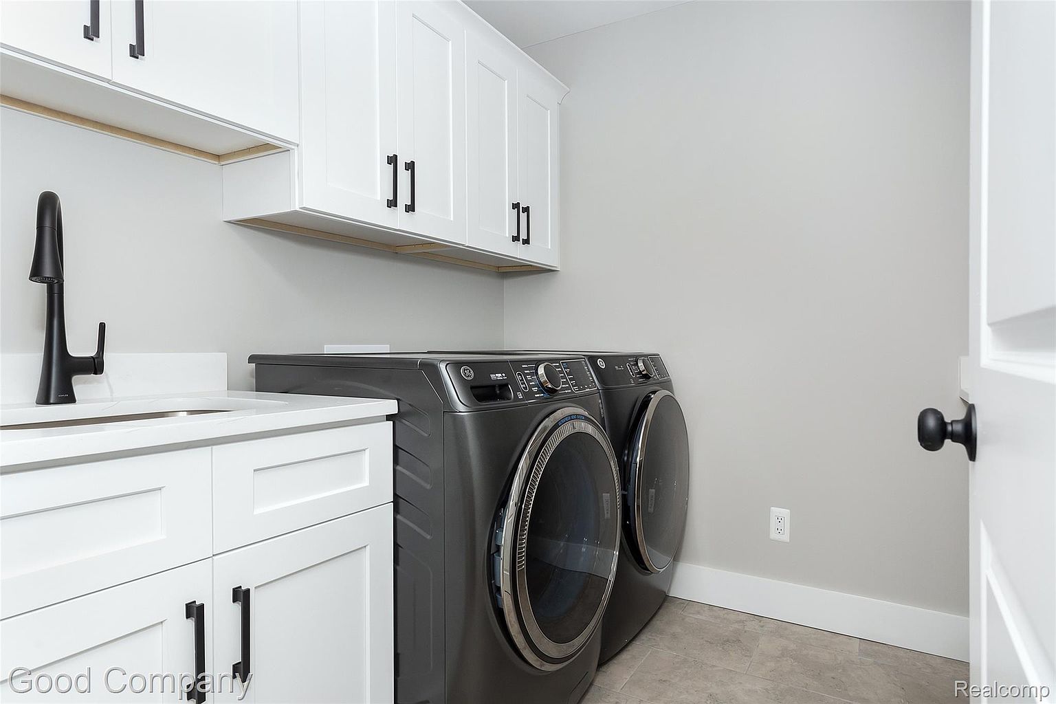 This is an interior shot of a laundry room featuring a modern washer and dryer set in a dark gray finish. The room has white cabinetry with black hardware, a white countertop with a sink, and light gray walls. The perspective is from a medium distance, showcasing the appliances and the overall layout of the laundry area.