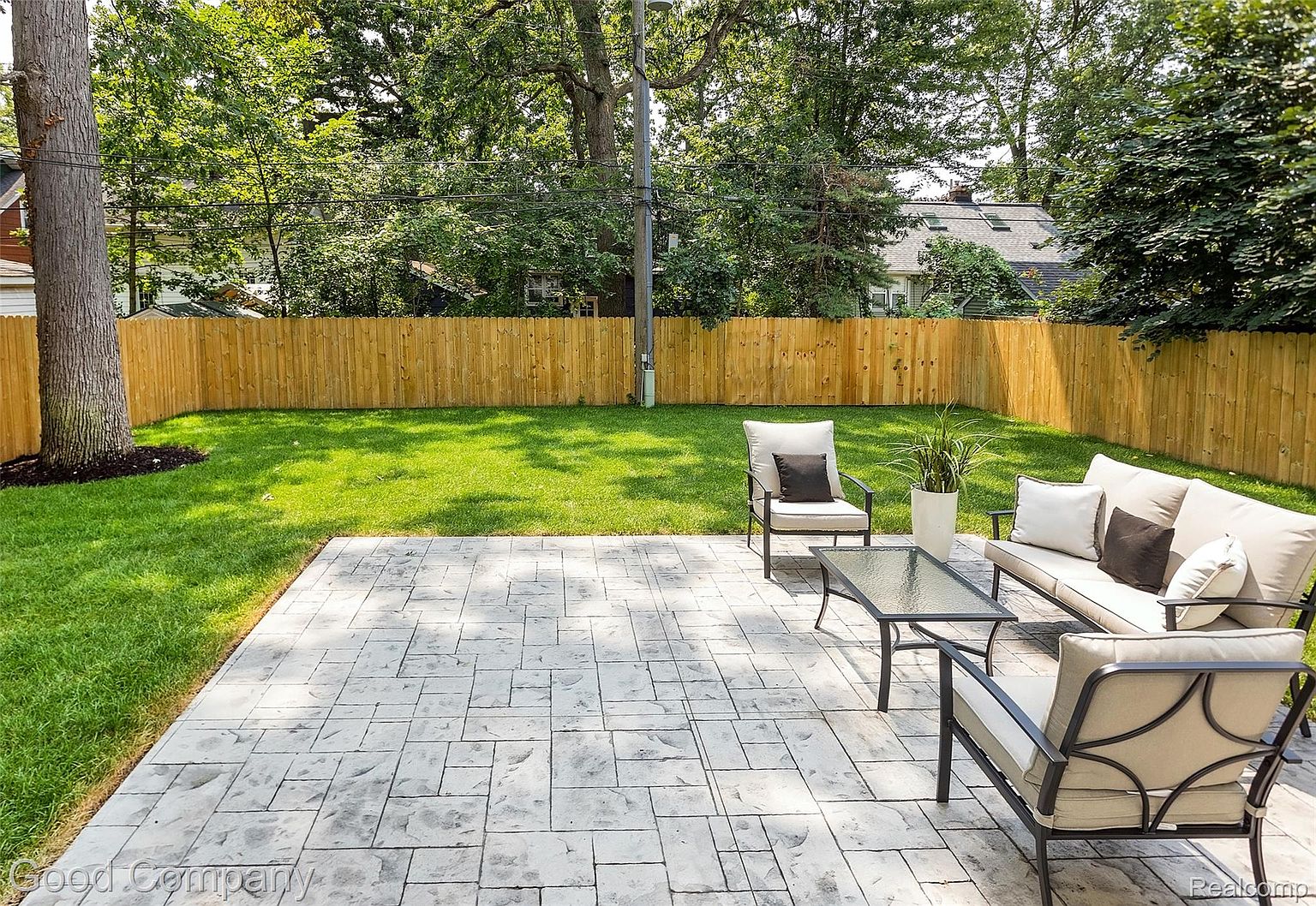 This image showcases a well-maintained backyard featuring a stone patio with outdoor seating, a lush green lawn, and a wooden fence providing privacy. The patio furniture includes a sofa, chairs, and a coffee table, creating an inviting outdoor living space. Mature trees surround the yard, adding to the serene and private atmosphere.