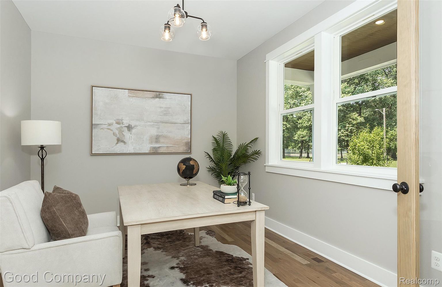 This is an interior shot of a home office or study. The room features a light-colored desk with a globe, books, and an hourglass, a comfortable white armchair with a brown pillow, and a framed abstract painting on the wall. Natural light streams in through a large window, creating a bright and inviting workspace.