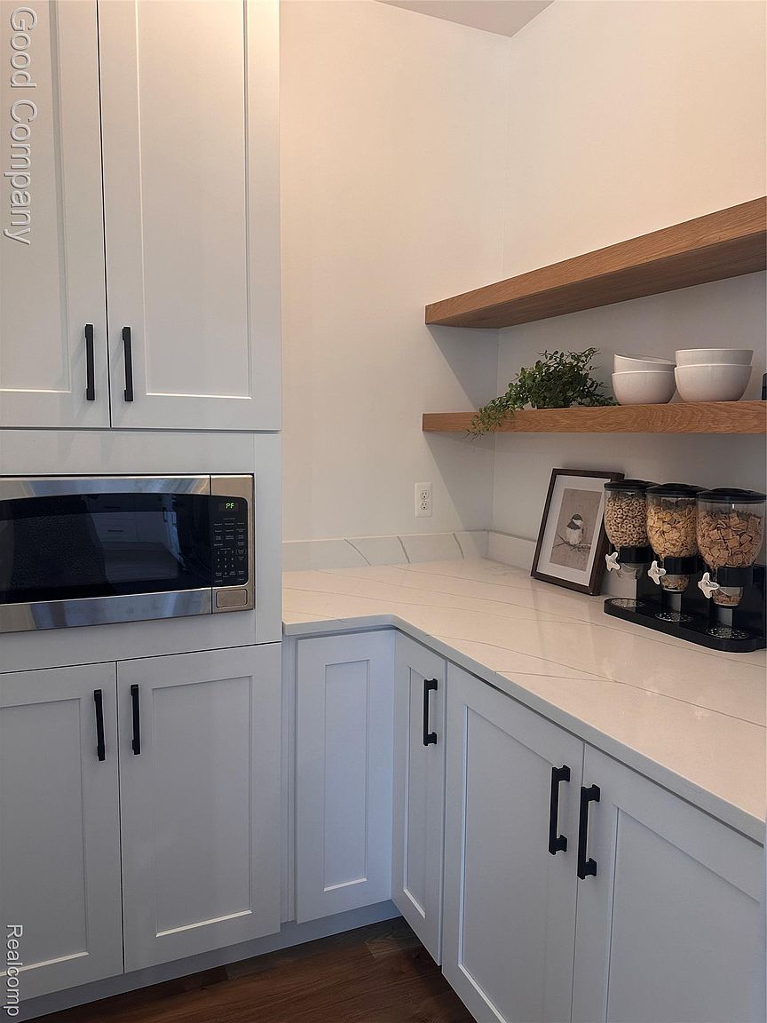 This is an interior shot of a well-organized pantry. The pantry features white cabinets with black hardware, a stainless steel microwave, light countertops, and open wooden shelves. A cereal dispenser and framed artwork add a touch of personality to the space.