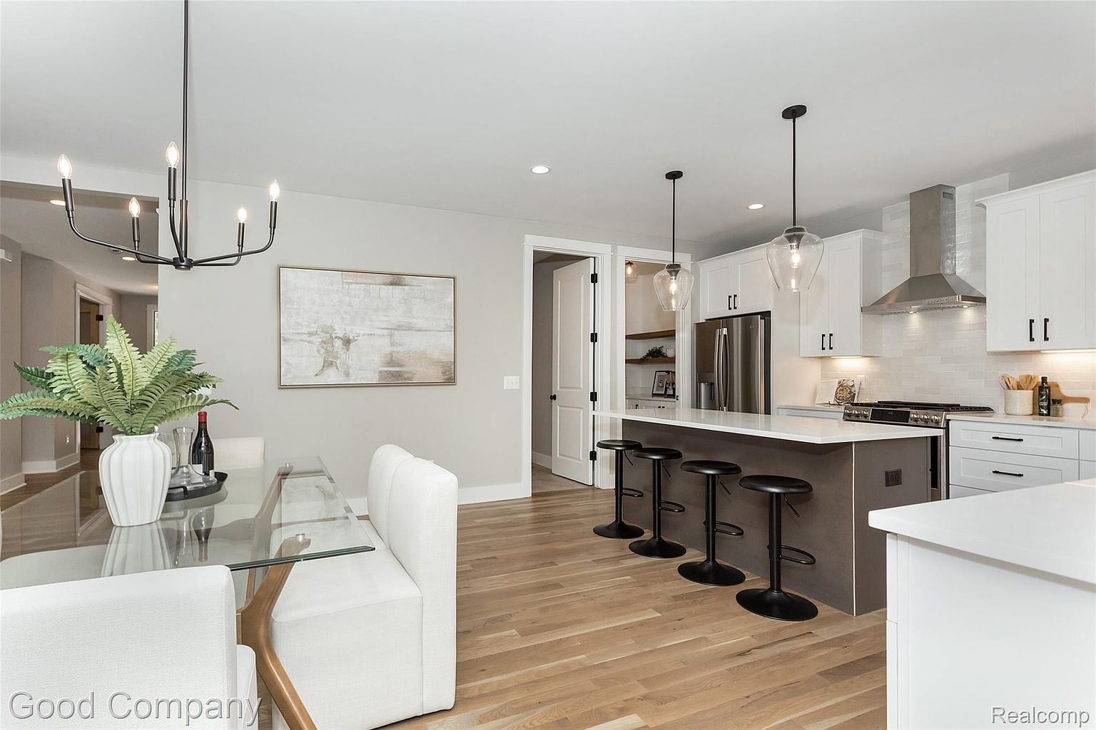 This interior shot showcases a modern kitchen and dining area. The kitchen features white cabinetry, stainless steel appliances, a kitchen island with bar seating, and pendant lighting. The dining area includes a glass-top table with white chairs and a bench, complemented by a modern chandelier and artwork, creating a bright and inviting space.