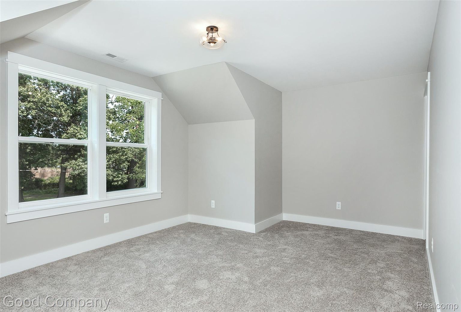 This is a well-lit, neutral-toned bedroom featuring gray walls, white trim, and gray carpet. A double window provides natural light and a view of trees outside. The room has a clean and simple design, making it a versatile space for various uses.