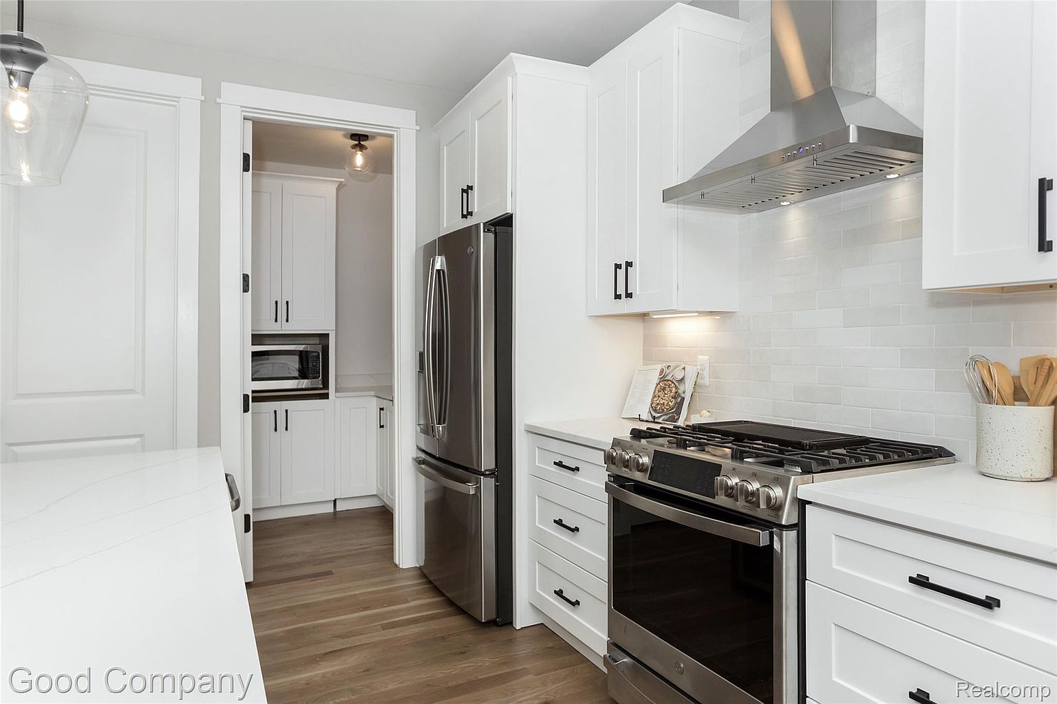 This is a bright and modern kitchen featuring white cabinetry, stainless steel appliances, and light wood flooring. The kitchen is well-lit, with a stainless steel range hood above the stove and a pantry visible through an open doorway. The overall impression is clean, stylish, and functional.