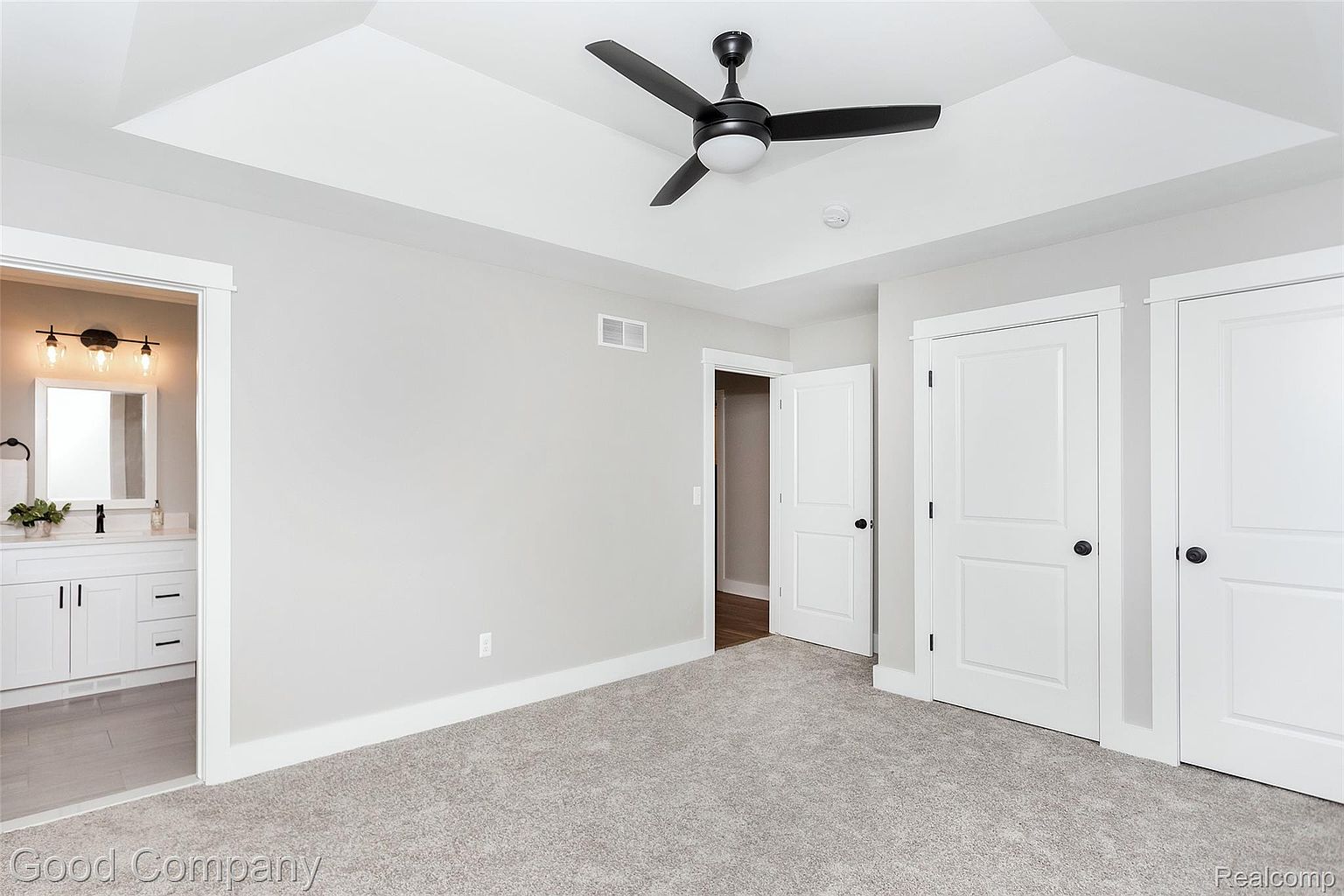 This is a primary bedroom featuring neutral gray walls and carpet. The room has a tray ceiling with a black ceiling fan. There are two white doors, one leading to a bathroom and another to a closet, adding to the functionality of the space.