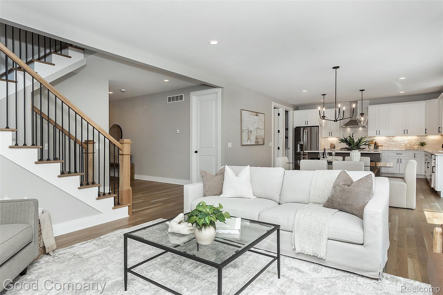 This is an interior shot of a living room that seamlessly transitions into a modern kitchen and dining area. The living room features a comfortable white sofa, a glass-topped coffee table, and a staircase with black iron railings. The open-concept design creates a spacious and inviting atmosphere, perfect for entertaining and family living.