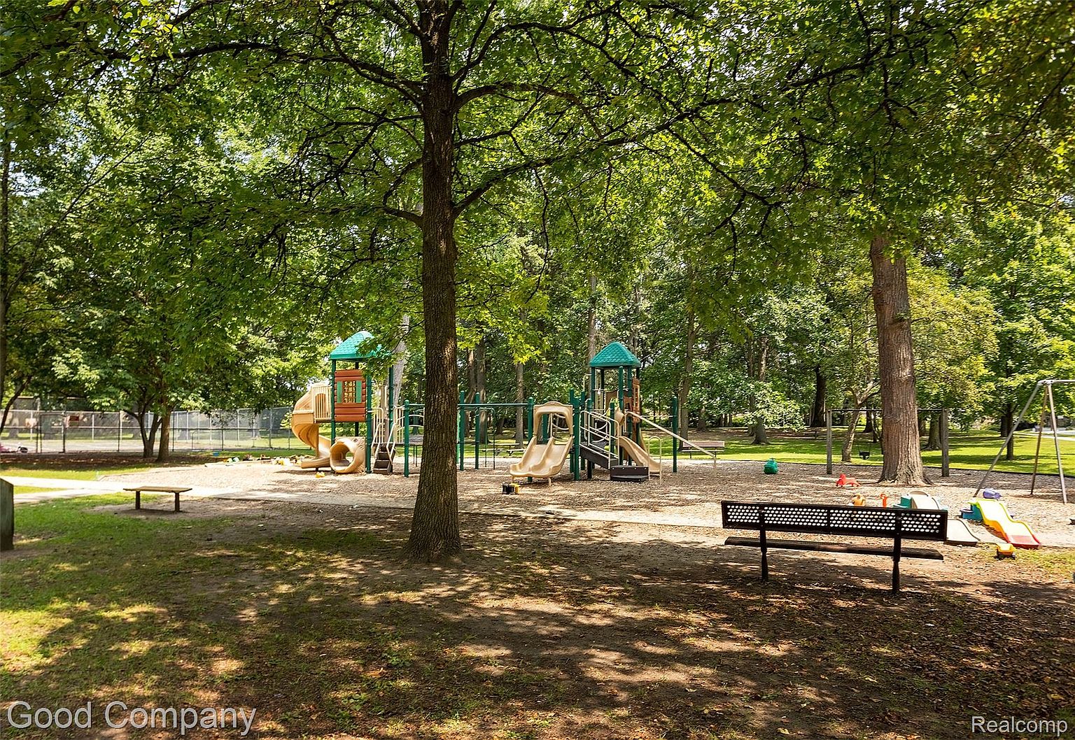 This image showcases a well-maintained playground area, likely part of a community park or residential complex. The playground features multiple play structures with slides, climbing areas, and swings, all set on a soft, sandy surface. Benches are strategically placed around the playground, offering seating for parents or guardians, and mature trees provide ample shade, creating a welcoming and safe environment for children.