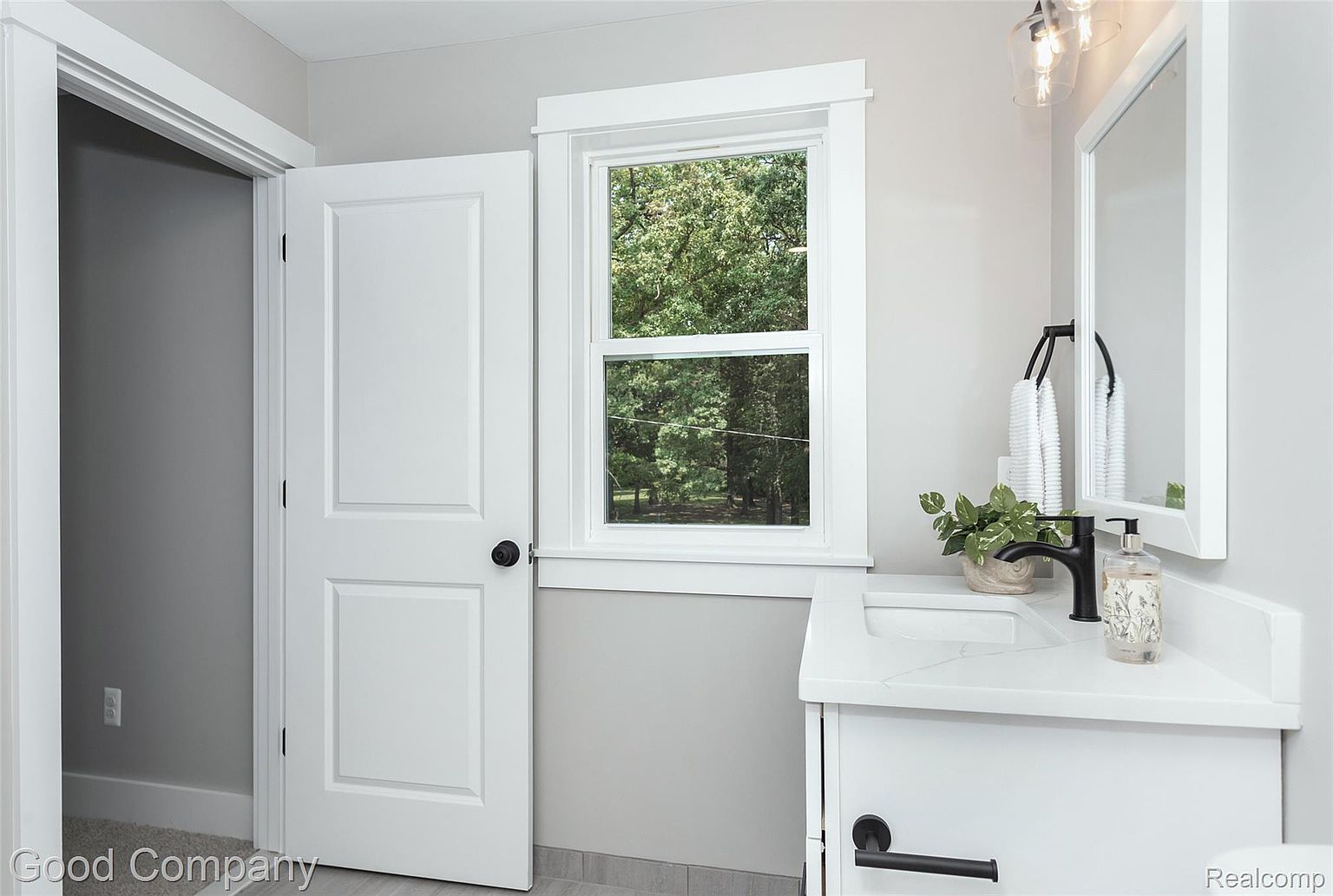 This is a bright and clean guest bathroom featuring a white vanity with a modern black faucet and hardware. A window provides natural light and a view of the outdoors, while a white-framed mirror hangs above the sink. The walls are painted a neutral gray, and the overall impression is fresh and inviting.
