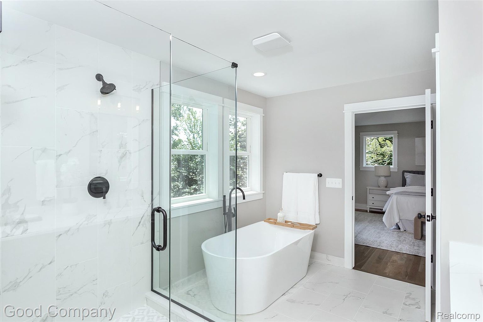 This is a bright and modern primary bathroom featuring a glass-enclosed shower with matte black fixtures and a freestanding soaking tub next to a window. The walls and floors are covered in white marble-patterned tiles, creating a clean and luxurious aesthetic. An open doorway leads to the primary bedroom, enhancing the sense of spaciousness.