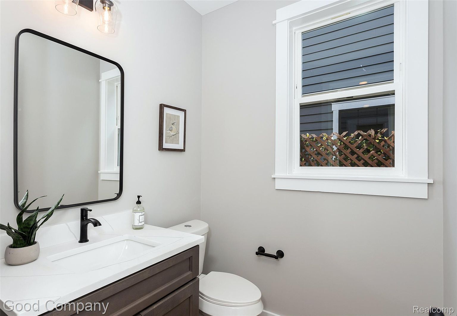 This is a well-lit guest bathroom featuring a modern vanity with a dark wood cabinet and a white countertop. A black framed mirror hangs above the sink, complemented by a black faucet and hardware. A window provides natural light, and a framed picture adds a touch of decor to the neutral-toned walls.
