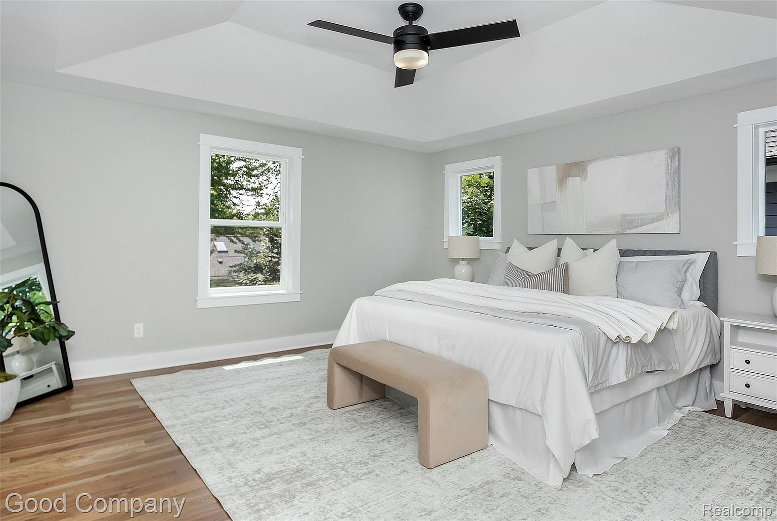This is a bright and airy primary bedroom featuring a large bed with white linens, a neutral-toned bench, and a light gray rug. Natural light streams in through the windows, complementing the soft gray walls and creating a serene atmosphere. A ceiling fan adds a modern touch.