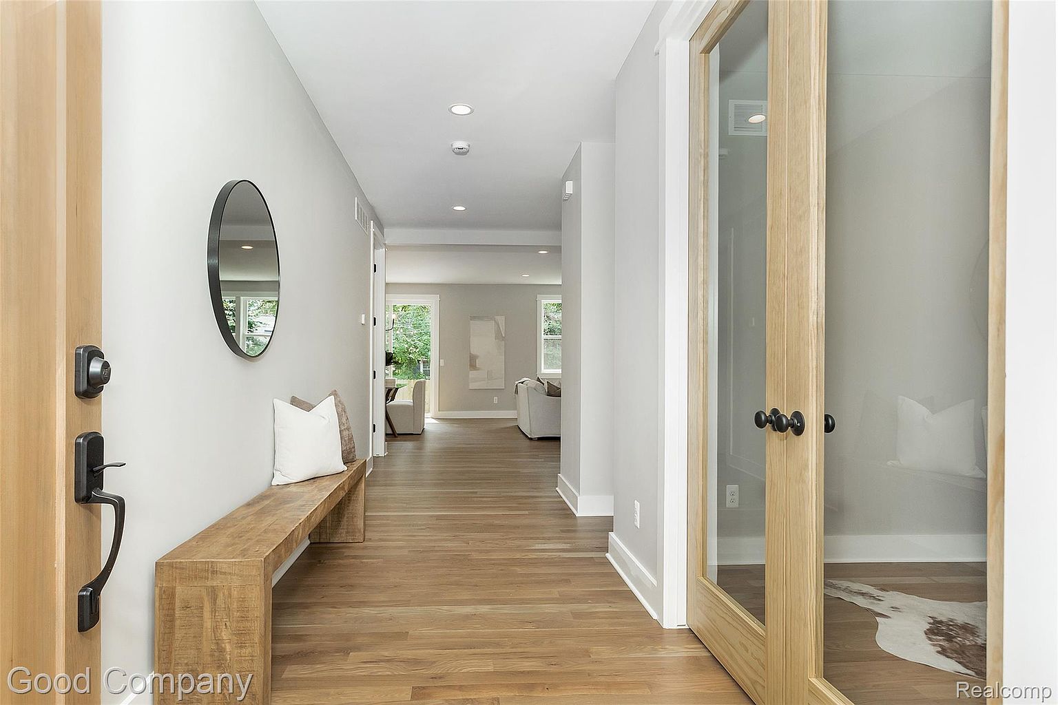This interior shot showcases a well-lit hallway with hardwood flooring, a wooden bench with pillows, and a round mirror on the wall. The hallway leads to a living area visible in the background, and a glass-paneled door is featured on the right. The perspective is from the entryway, creating a welcoming and stylish impression.