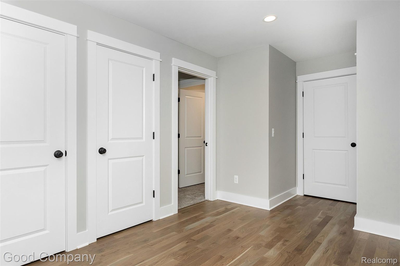 This interior shot showcases a hallway with light gray walls and light wood flooring. Three white doors with black hardware are visible, adding a clean and modern aesthetic. The hallway leads to another room, offering a glimpse of a carpeted area and another white door.