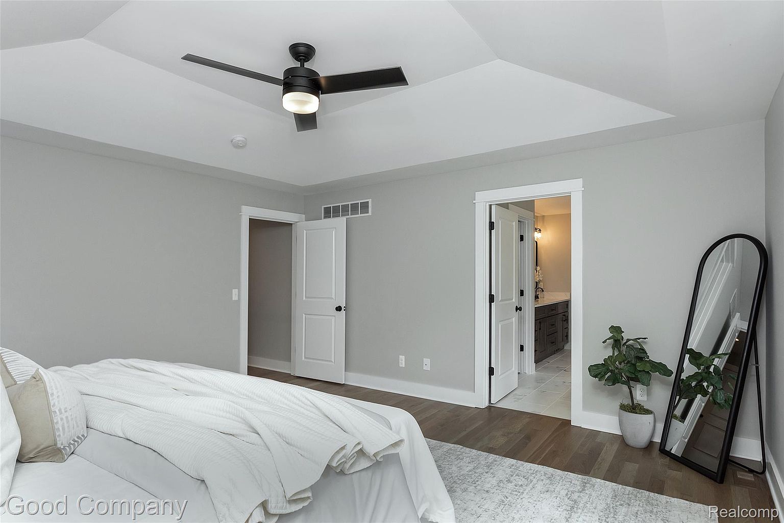 This is a primary bedroom featuring a tray ceiling with a black ceiling fan. The walls are painted a neutral gray, and there are two white doors, one leading to another room and the other to a bathroom. A full-length mirror and a potted plant add to the decor, and a bed with white linens is partially visible in the foreground.