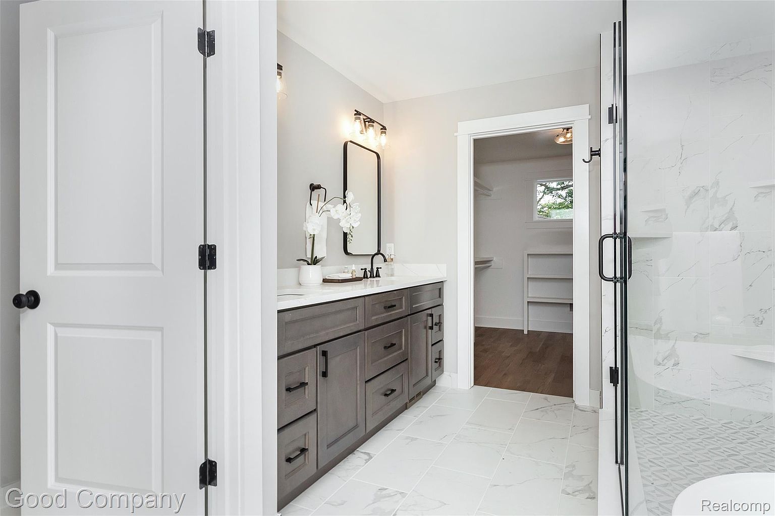 This is a well-lit primary bathroom featuring a gray vanity with a white countertop, a black framed mirror, and modern lighting fixtures. The floor is covered in white marble-like tiles, and a glass-enclosed shower is visible on the right. An open doorway leads to a walk-in closet, adding to the bathroom's spacious feel.