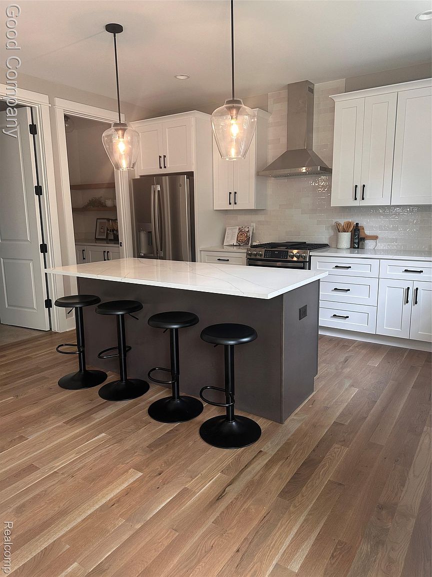 This is a well-lit kitchen featuring a central island with a white countertop and dark base, surrounded by four black bar stools. The kitchen includes white cabinets with black hardware, stainless steel appliances, and pendant lighting above the island. The hardwood flooring adds warmth to the space, creating a modern and inviting atmosphere.