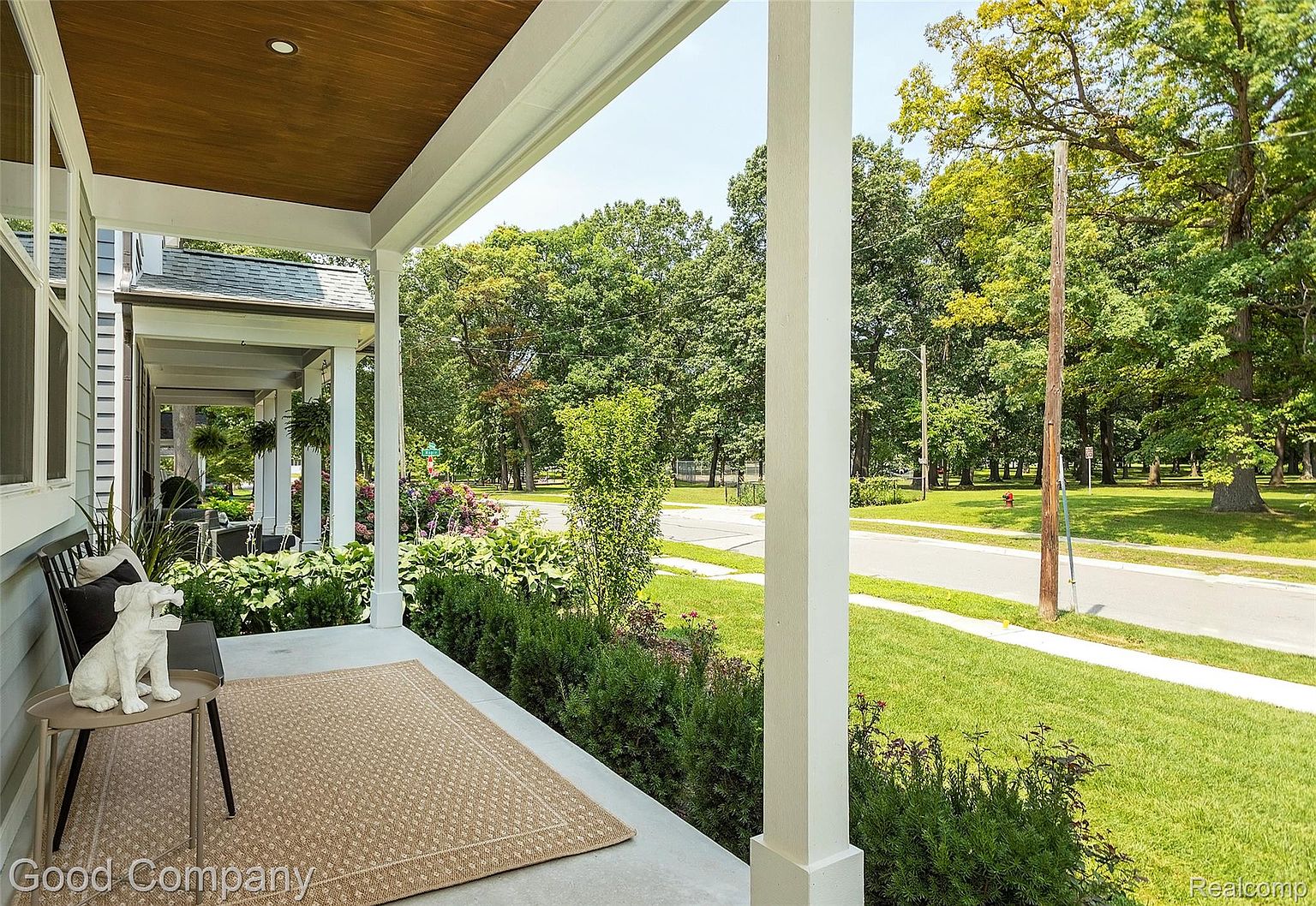 This image showcases a charming porch area, featuring a neutral-toned rug, a small table with a decorative dog statue, and a bench. The porch is framed by white columns and offers a view of a well-maintained lawn, lush greenery, and a quiet street. The overall impression is inviting and serene, highlighting the home's curb appeal and outdoor living space.