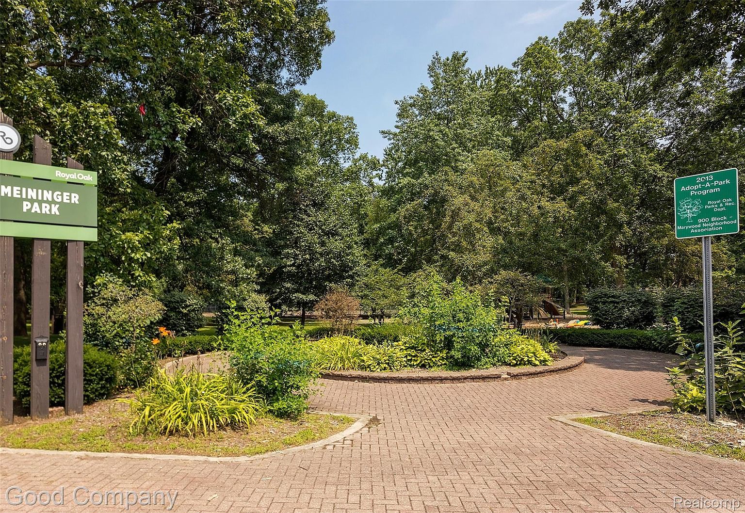 This image showcases the entrance to Meininger Park in Royal Oak, featuring a brick pathway leading into the park. Lush greenery and mature trees surround the area, creating a welcoming and serene atmosphere. Signage indicates the park's name and participation in the Adopt-A-Park program, highlighting community involvement.
