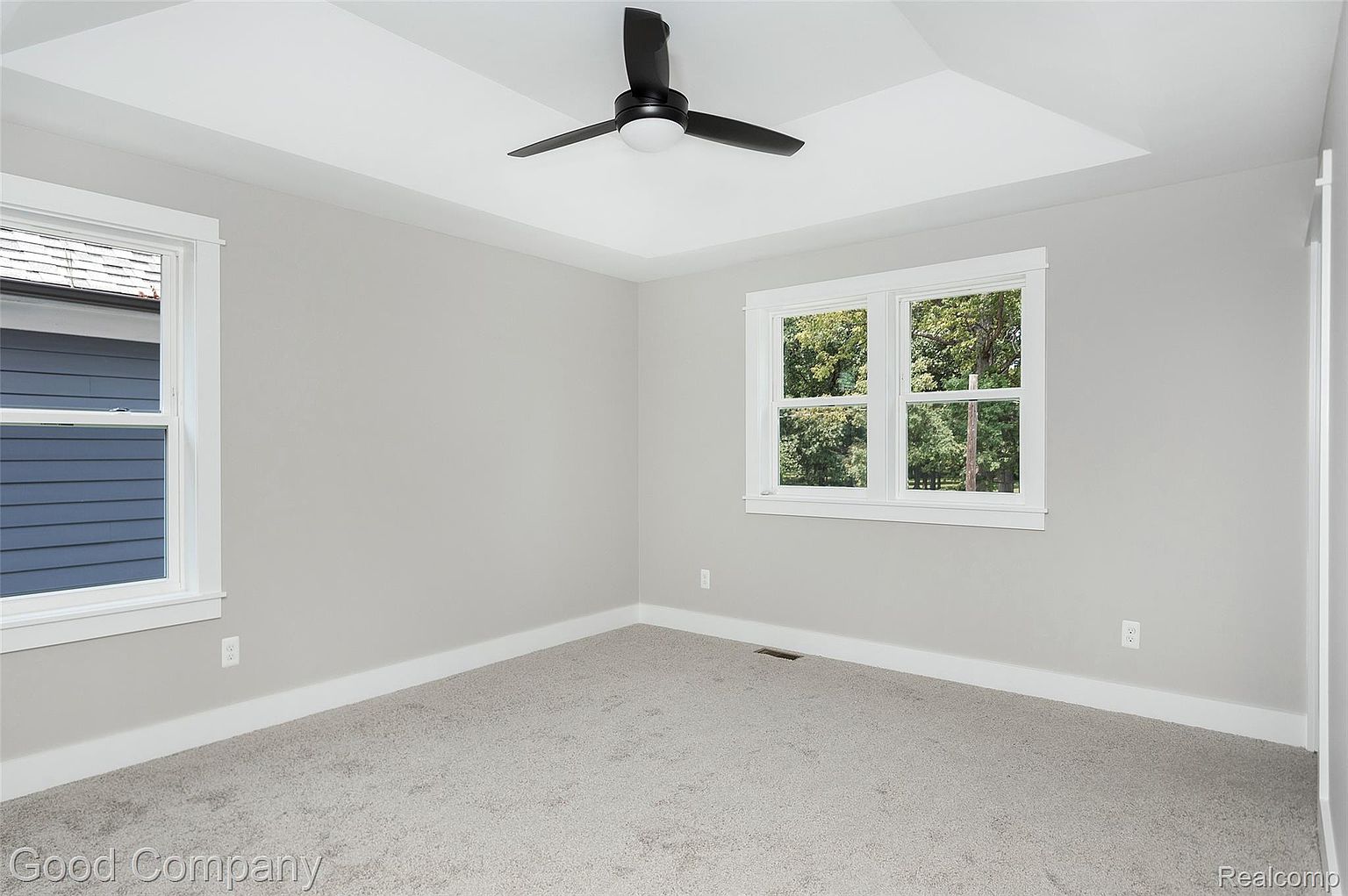 The image showcases a well-lit bedroom with neutral gray walls and light gray carpet. A ceiling fan with a light fixture is centered on the ceiling. Two windows provide natural light, one showing the exterior of the house and the other showing trees and greenery.