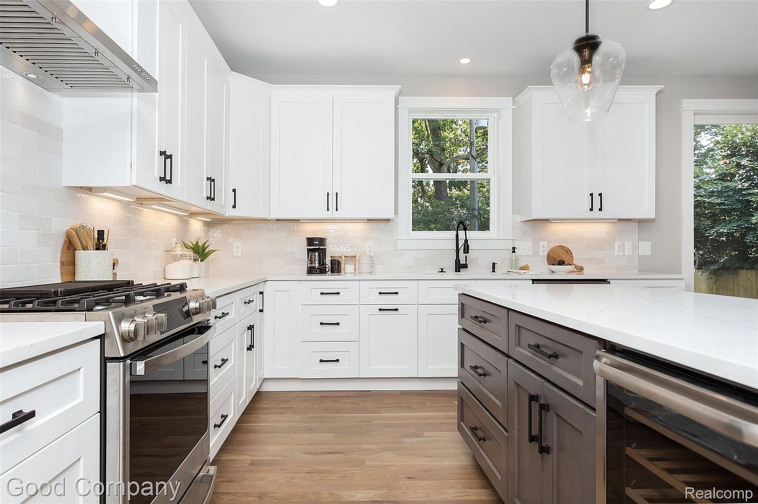 This is a bright and modern kitchen featuring white cabinets with black hardware, stainless steel appliances, and hardwood floors. A kitchen island with a contrasting gray base and white countertop provides additional workspace and storage. Natural light streams in through a window above the sink, creating a welcoming atmosphere.