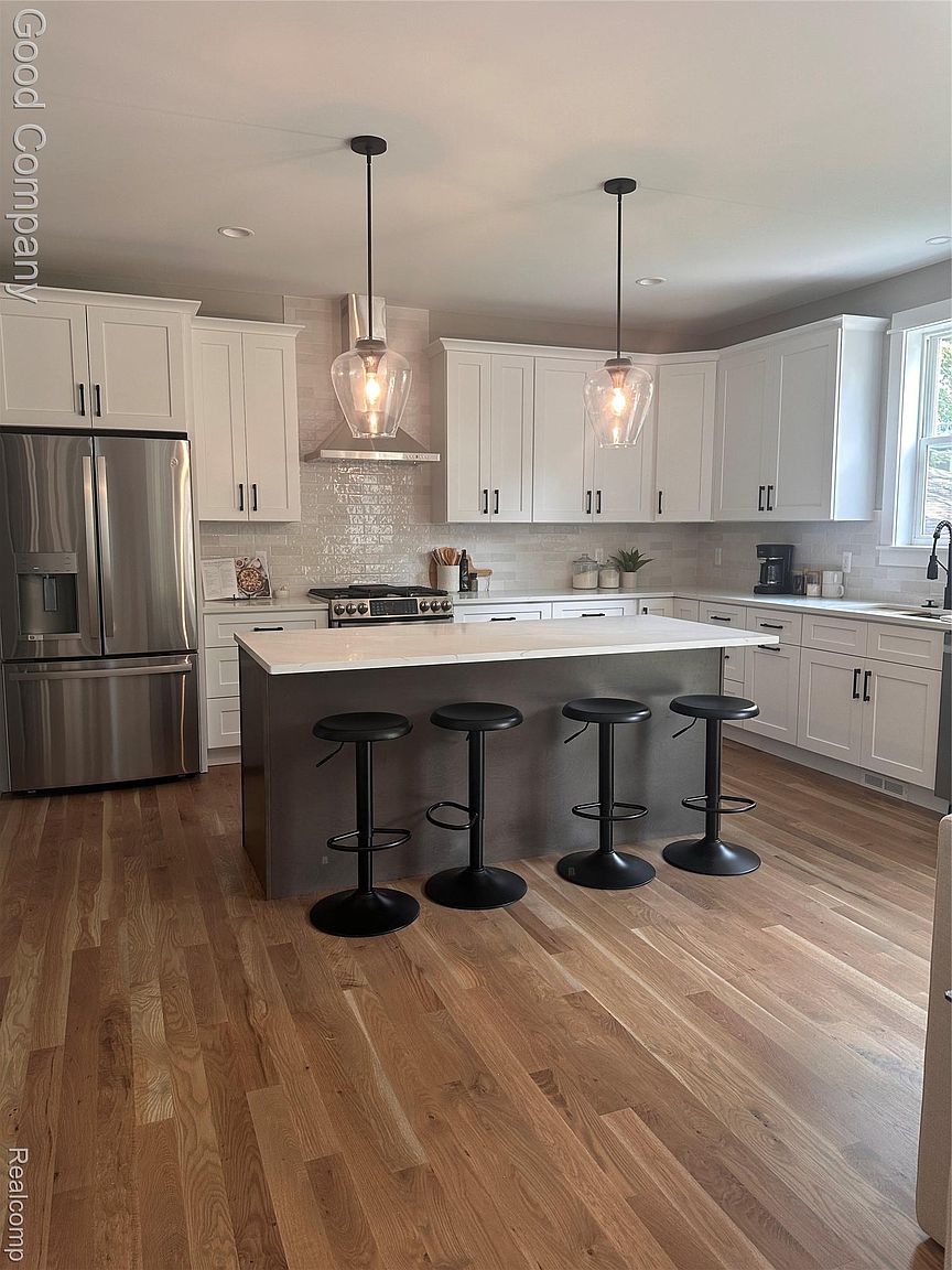 This is a well-lit kitchen featuring white cabinets with black hardware, stainless steel appliances, and a gray island with a white countertop. Four black bar stools are positioned around the island, and pendant lights hang above it. The hardwood floors add warmth to the space, creating a modern and inviting atmosphere.