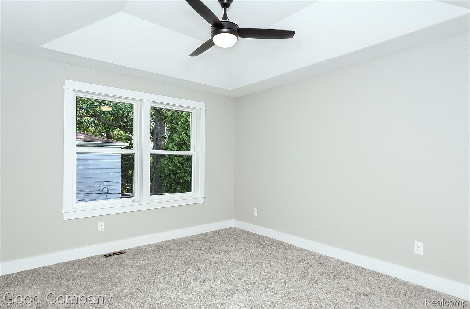 This is a bright and airy bedroom featuring neutral gray walls and carpet. A large window provides natural light and a view of the outdoors. A modern ceiling fan adds a touch of contemporary style to the room.