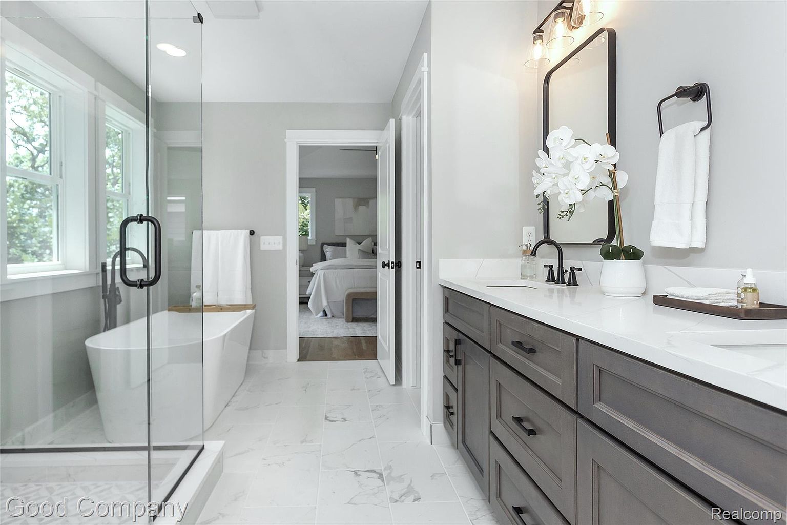 This is a primary bathroom featuring a modern design with a freestanding bathtub enclosed by a glass shower, a double vanity with gray cabinetry and white countertops, and marble-look tile flooring. The bathroom opens into a bedroom, creating a seamless and luxurious space. The overall impression is clean, bright, and sophisticated.