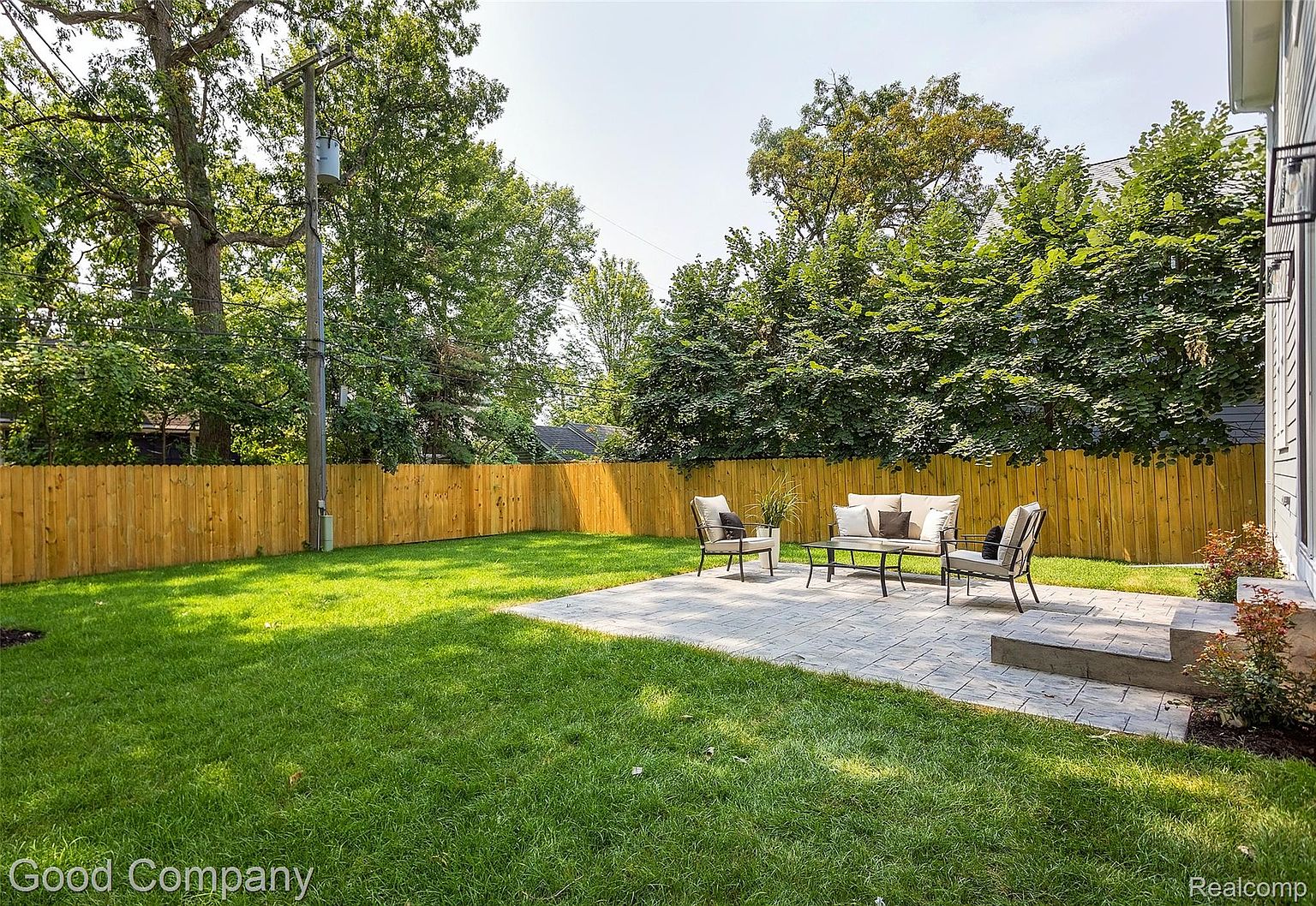 This image showcases a well-maintained backyard featuring a lush green lawn, a wooden fence, and a paved patio area with outdoor seating. The yard is surrounded by mature trees, providing privacy and shade. The overall impression is a serene and inviting outdoor space, perfect for relaxation and entertaining.