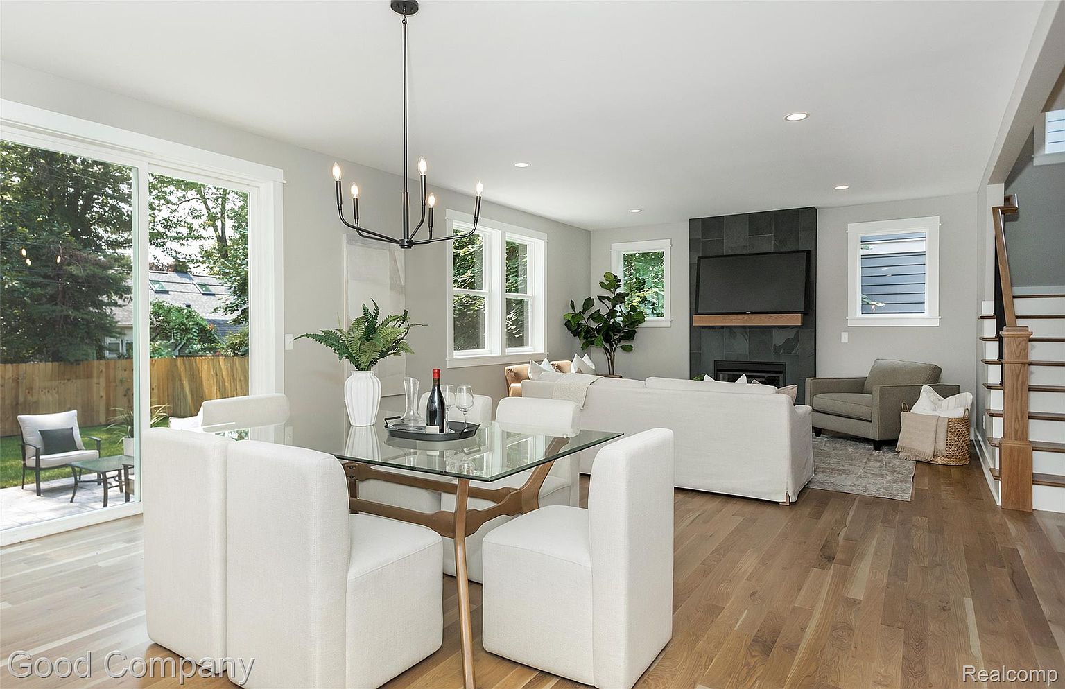 This is an interior shot of a dining room and living room area. The dining area features a glass-top table with white upholstered chairs, illuminated by a modern chandelier. The living room has a fireplace with a dark stone surround and a large television above, complemented by neutral-toned furniture and hardwood floors, creating a bright and inviting space.