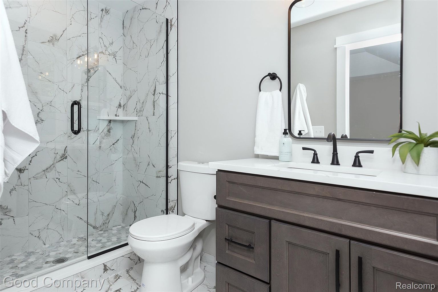 This is a well-lit bathroom featuring a glass-enclosed shower with marble-patterned walls and a dark-framed door. A white toilet sits next to a vanity with a dark wood cabinet, a white countertop, and a black faucet. The mirror above the sink has a matching black frame, and the overall aesthetic is modern and clean.