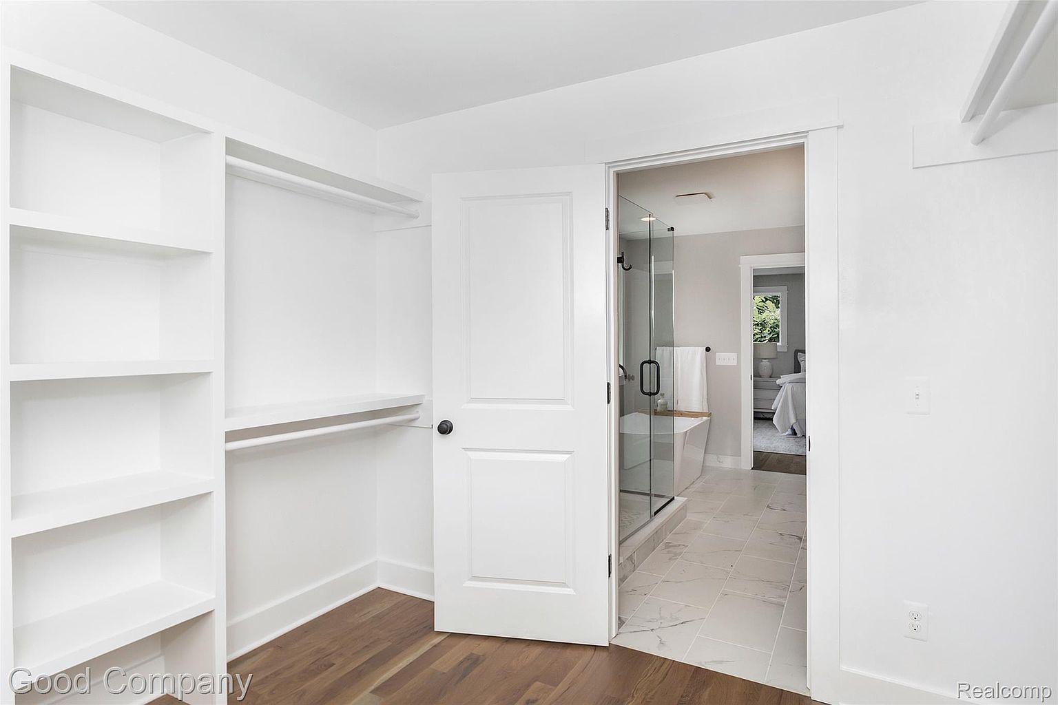 This image showcases a well-organized walk-in closet with custom white shelving and hanging rods. The closet opens into a bright bathroom with a glass-enclosed shower and a glimpse of the bedroom beyond. The hardwood flooring adds warmth to the space, creating a clean and inviting atmosphere.