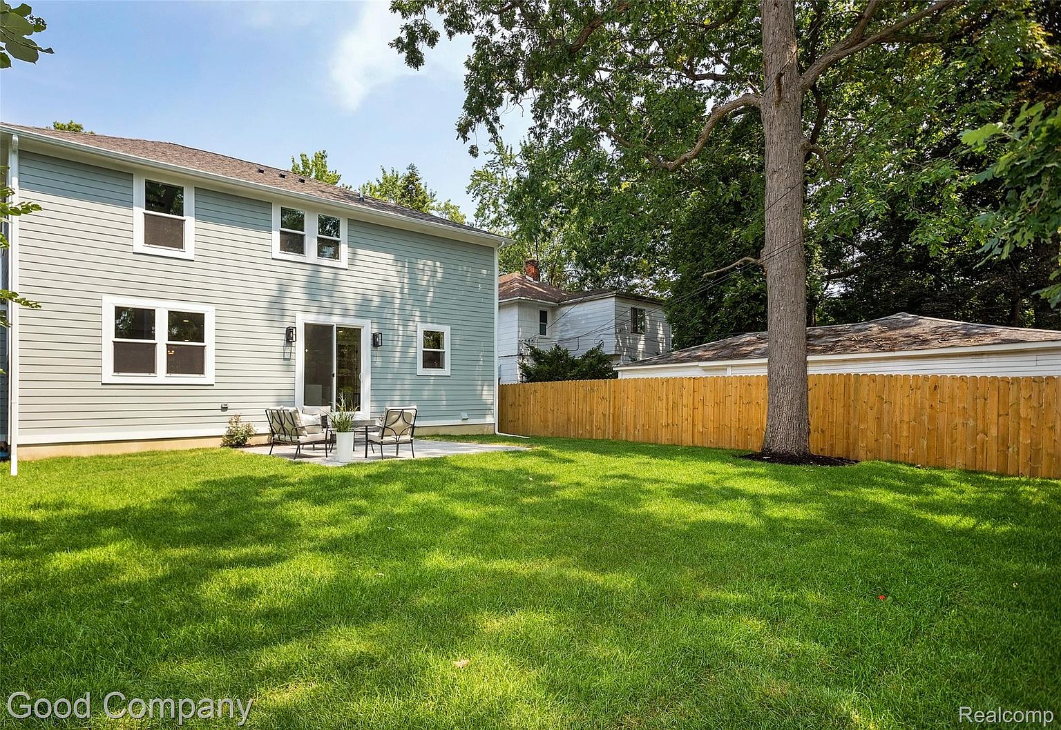 This image showcases a well-maintained backyard featuring a lush green lawn, a light blue two-story house with white trim, and a patio area with outdoor seating. A wooden fence provides privacy, and mature trees add shade and character to the property. The overall impression is a serene and inviting outdoor space.