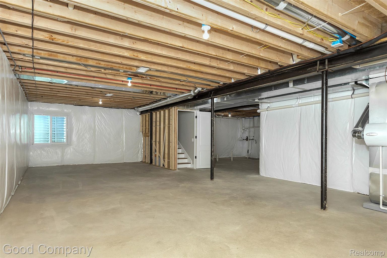 This is an unfinished basement with exposed wooden ceiling beams and a concrete floor. The walls are partially covered with white plastic sheeting, and there's a framed doorway leading to a staircase. The space is well-lit, giving a sense of potential for customization and expansion.