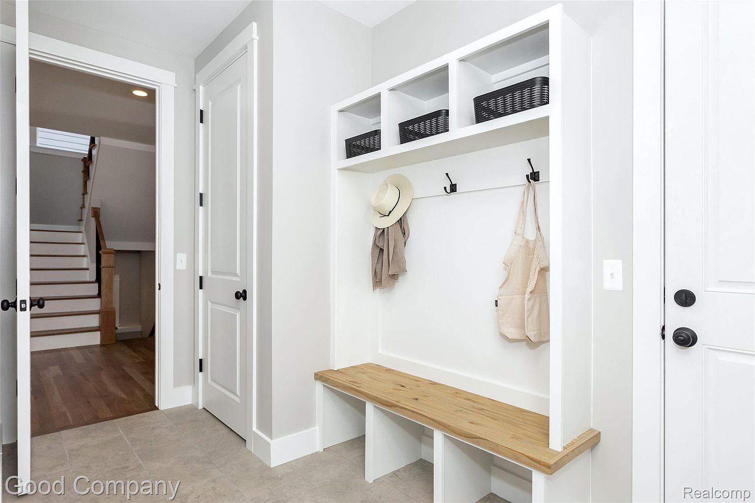 This interior shot showcases a well-organized mudroom area with built-in storage, including cubbies, hooks, and a wooden bench. The neutral color palette and clean lines contribute to a modern and inviting entryway. An open doorway reveals a staircase, adding depth to the image.