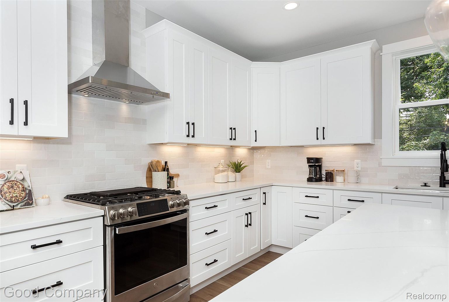This is a well-lit kitchen featuring white cabinetry with black hardware, stainless steel appliances, and light countertops. The kitchen includes a stainless steel range hood and a window providing natural light. The overall impression is clean, modern, and functional.