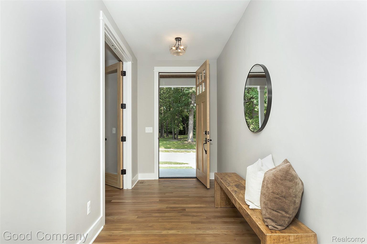 This interior shot showcases a bright and inviting hallway with hardwood flooring and light gray walls. A wooden bench with decorative pillows sits against the wall, complemented by a round mirror. An open doorway leads to the outside, providing a glimpse of greenery and natural light, while another door is slightly ajar on the left.