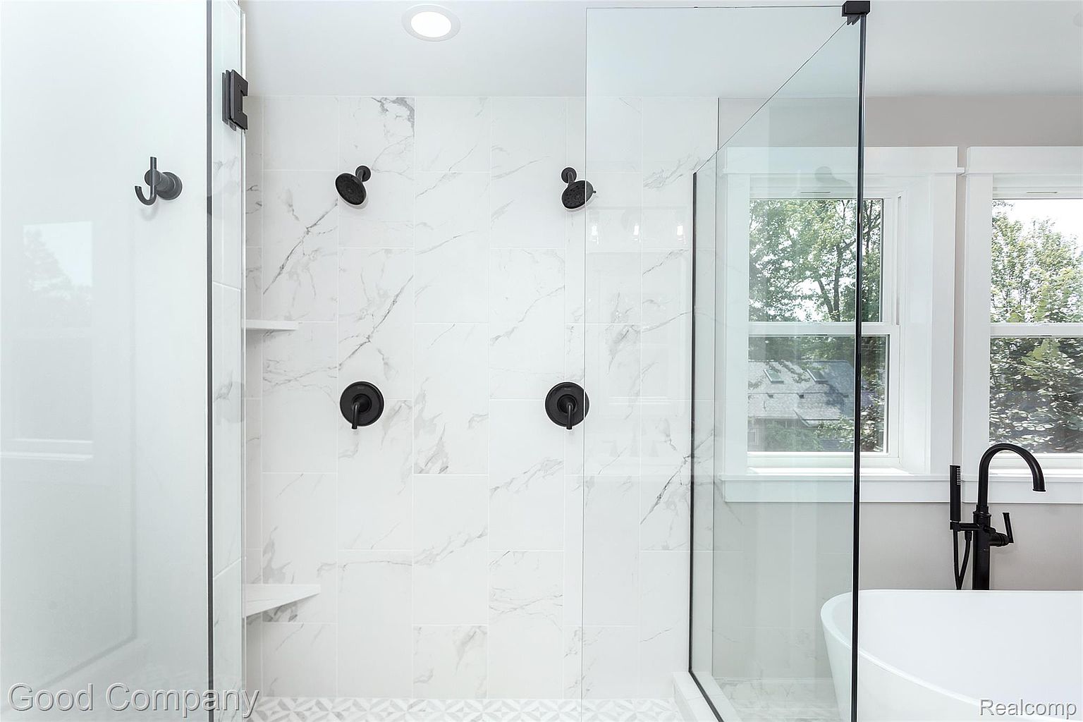 This is a modern primary bathroom featuring a glass-enclosed shower with white marble-patterned tile walls and matte black fixtures. The shower includes dual shower heads and built-in shelving. Natural light streams in through the windows, highlighting the clean and contemporary design.