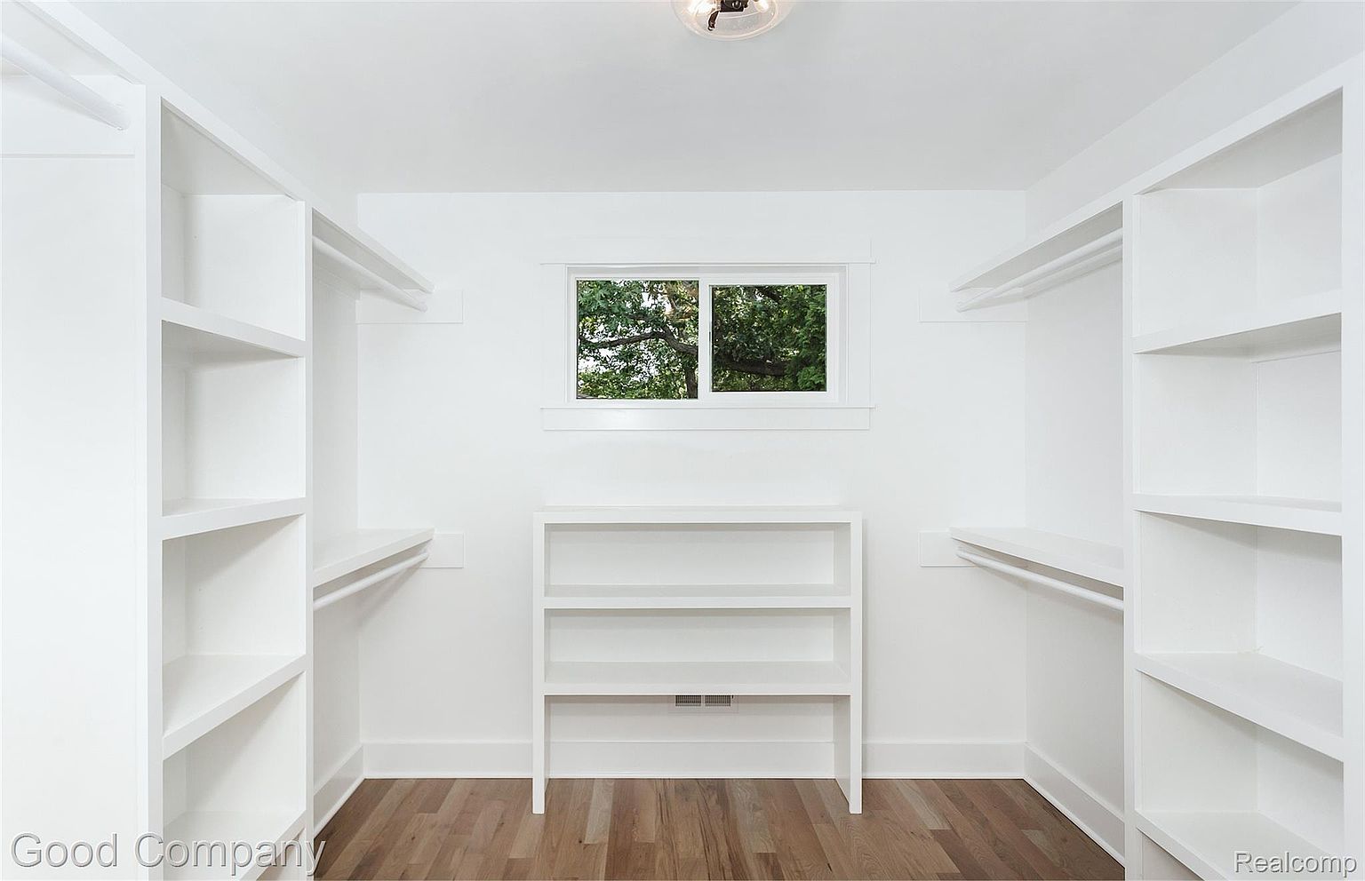 This is a bright and spacious walk-in closet featuring custom white shelving and hanging rods on both sides. A central shelving unit sits beneath a window, providing additional storage. The hardwood flooring adds a touch of warmth to the otherwise all-white space, creating a clean and organized aesthetic.