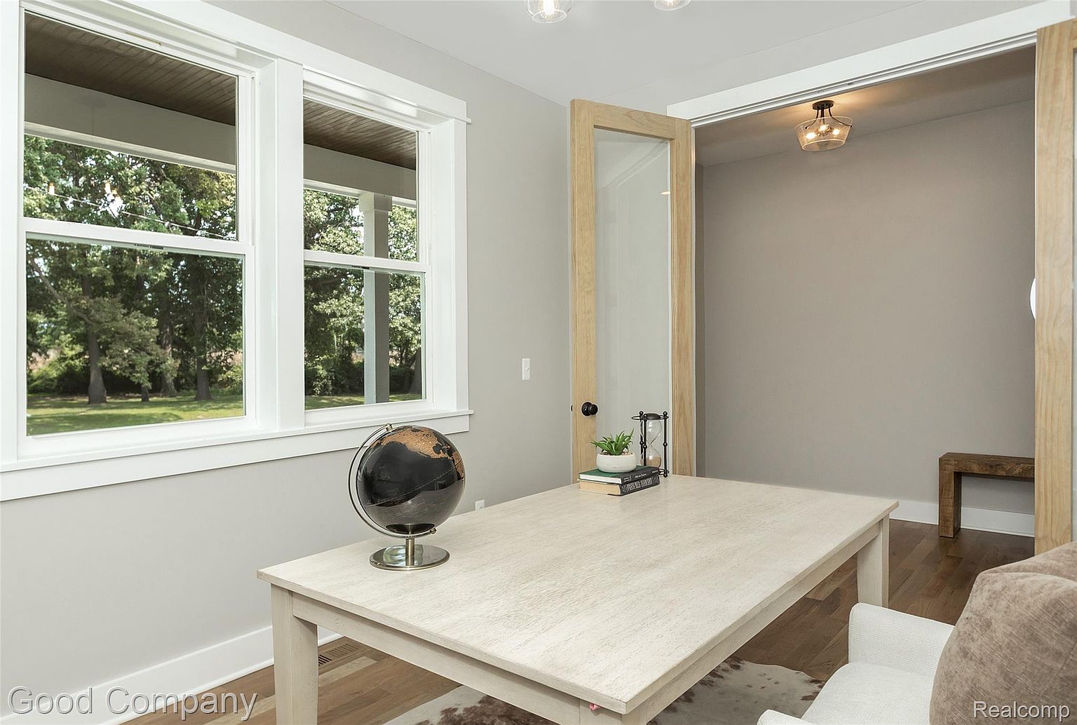This is an interior shot of a home office or study, featuring a light-colored wooden desk with a globe and decorative items. Natural light floods the room through a large window, and an open doorway leads to another room with a modern light fixture. The room is painted in a neutral gray tone, creating a calm and professional atmosphere.