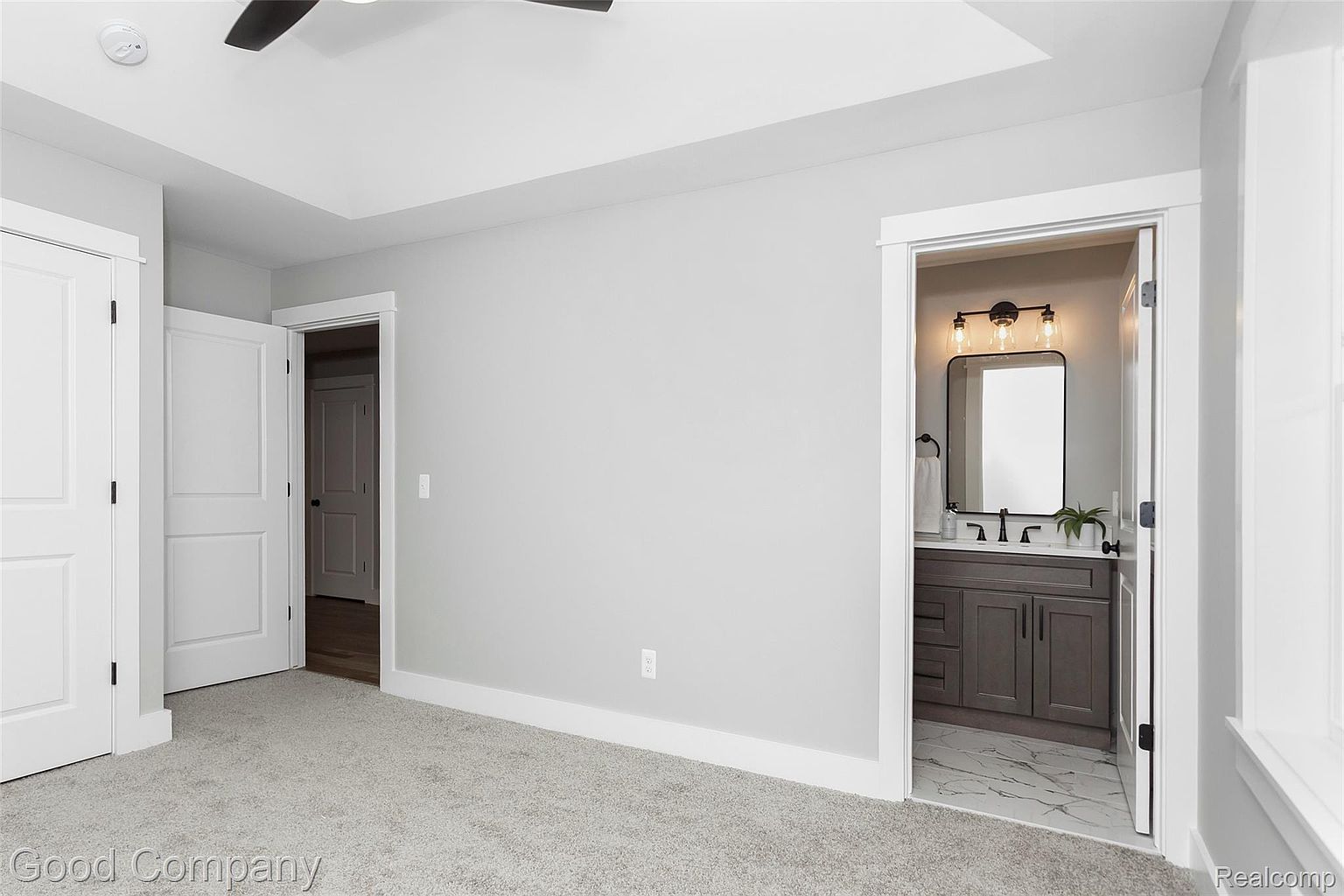 This is a primary bedroom featuring neutral gray walls and carpet. There are two white doors, one leading to a closet and the other to another room. An open doorway reveals a bathroom with a gray vanity, black fixtures, and marble-look flooring, creating a seamless transition between spaces.