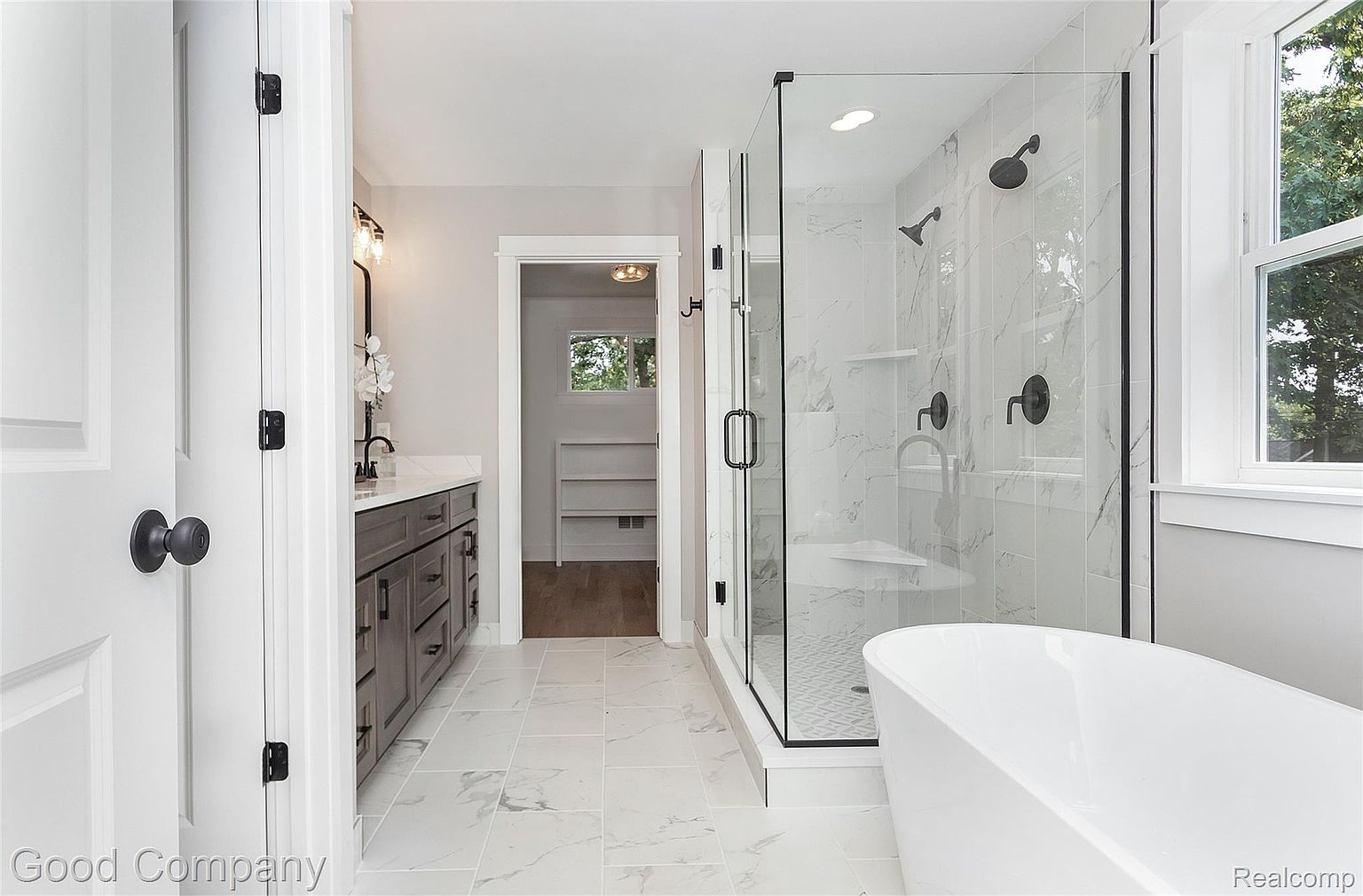 This is a bright and modern primary bathroom featuring a freestanding white bathtub, a glass-enclosed shower with black fixtures, and a gray vanity with a white countertop. The floor is covered in large marble-patterned tiles, and a doorway leads to a walk-in closet. The overall impression is clean, luxurious, and well-designed.