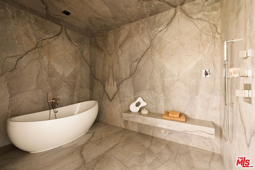This luxurious primary bathroom features a minimalist aesthetic with large-format, veined stone tiles covering the walls and floor. A sleek, freestanding white soaking tub is positioned on the left, while a floating stone bench provides a functional surface for towels and decor on the right. The space is equipped with a modern wall-mounted shower system, creating a spa-like atmosphere with a cohesive, high-end design.