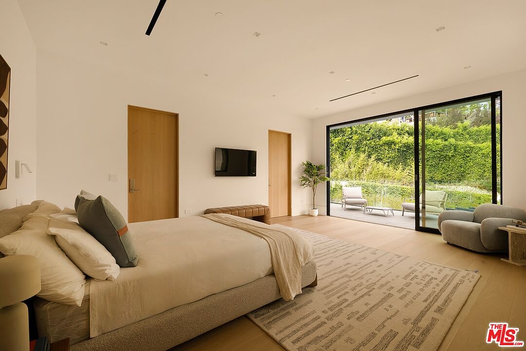 This spacious primary bedroom features a minimalist aesthetic with light wood flooring and neutral-toned walls. A large bed with a textured rug serves as the focal point, while floor-to-ceiling sliding glass doors provide seamless access to an outdoor patio overlooking lush greenery. The room is accented by a wall-mounted television, a low-profile bench, and a modern armchair, creating a serene and sophisticated retreat.